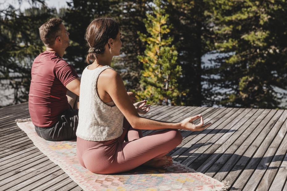 Two people meditating on a wooden deck beside a tree-lined lake
